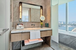 Un baño moderno con encimera de mármol, un espejo grande con luz redonda y vistas a un paisaje urbano a través de un ventanal.