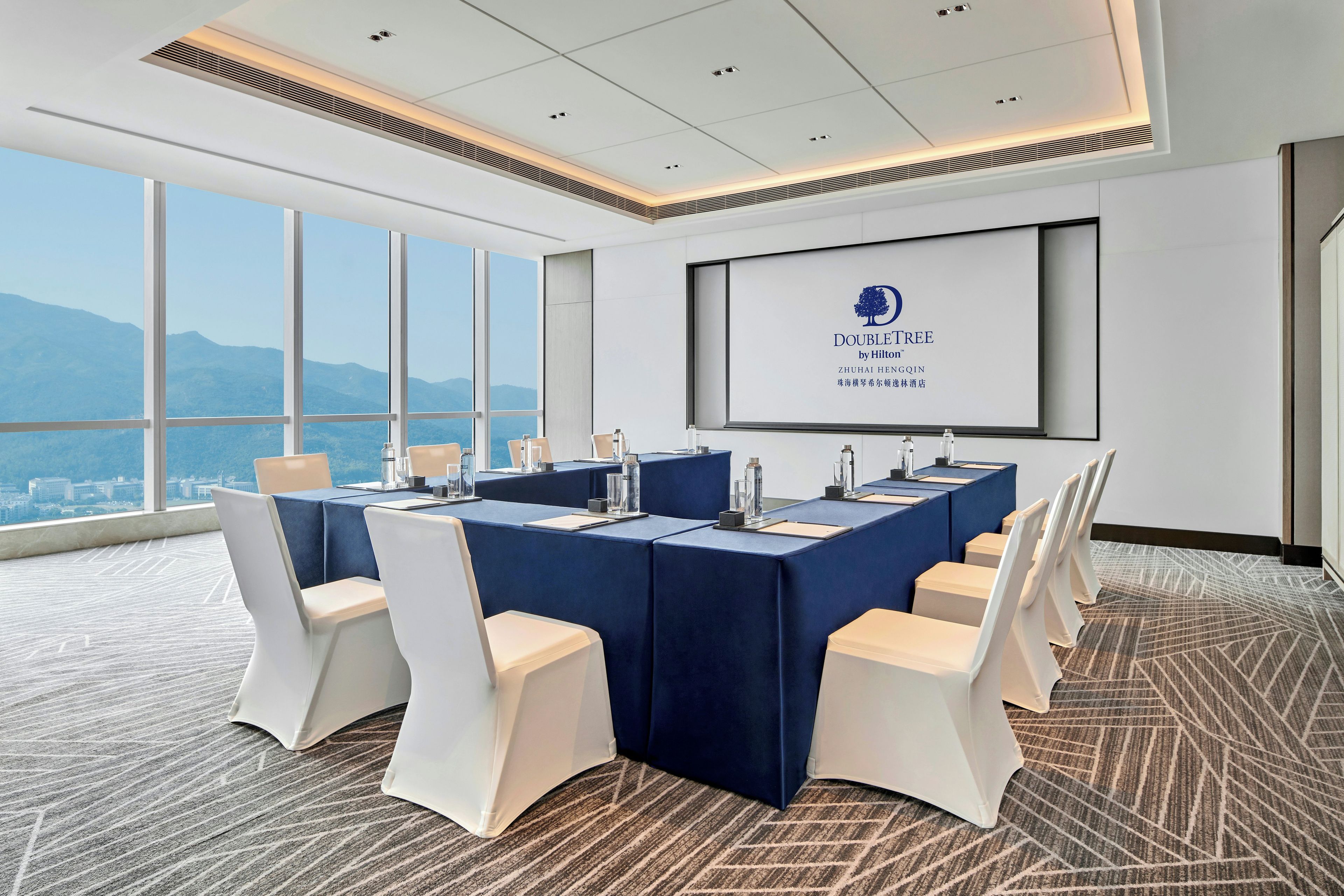 Un salón de conferencias con una pantalla grande que muestra el logotipo de DoubleTree by Hilton, una vista panorámica de la ciudad a través de amplias ventanas y un piso alfombrado.