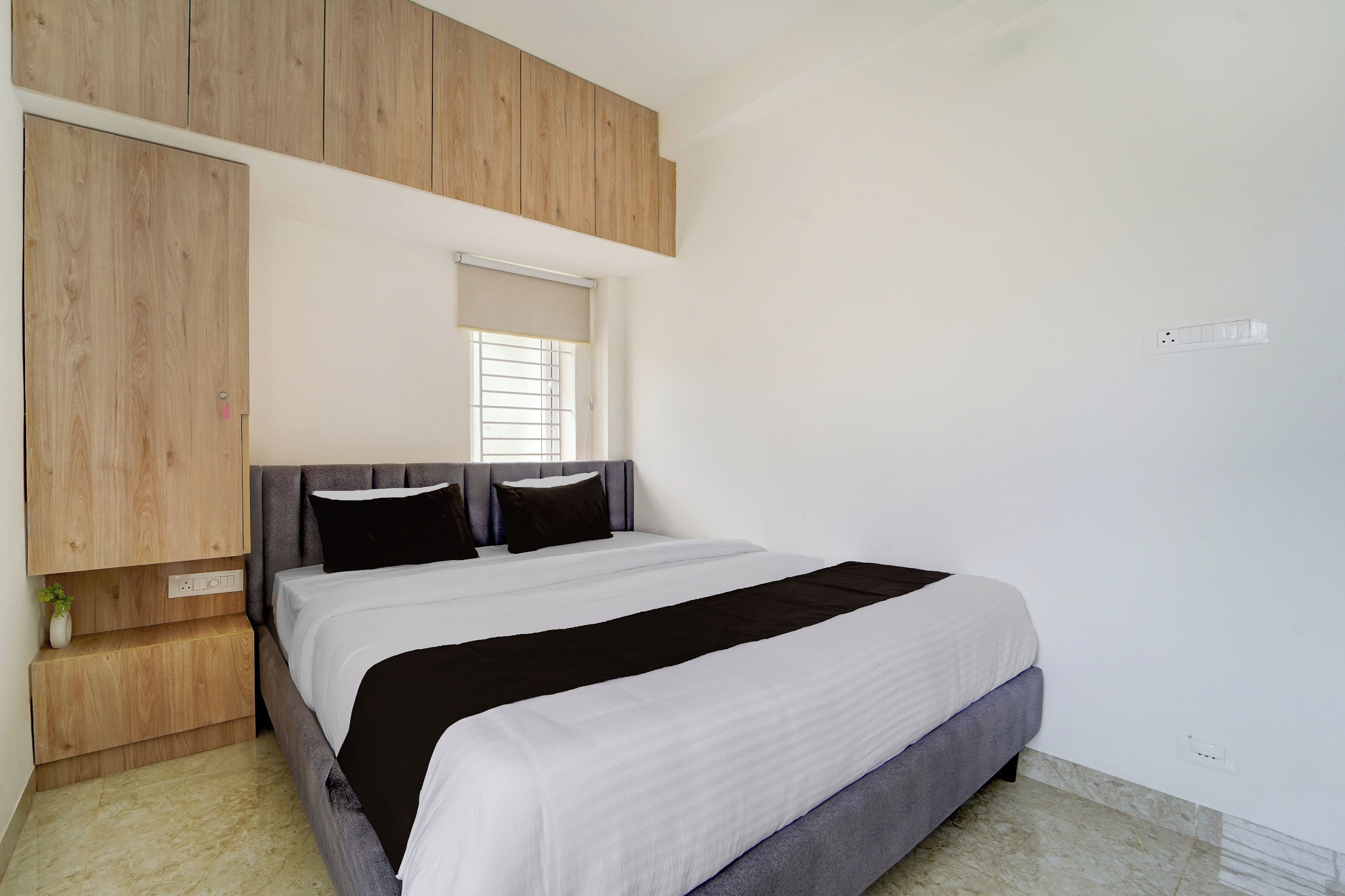 Un dormitorio con una cama, una ventana con persianas, paneles de madera sobre la cama y una pequeña planta en la mesita de noche.