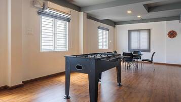 Una habitación con mesa de futbolín, dos ventanas con persianas, ventilador de techo y dardo.