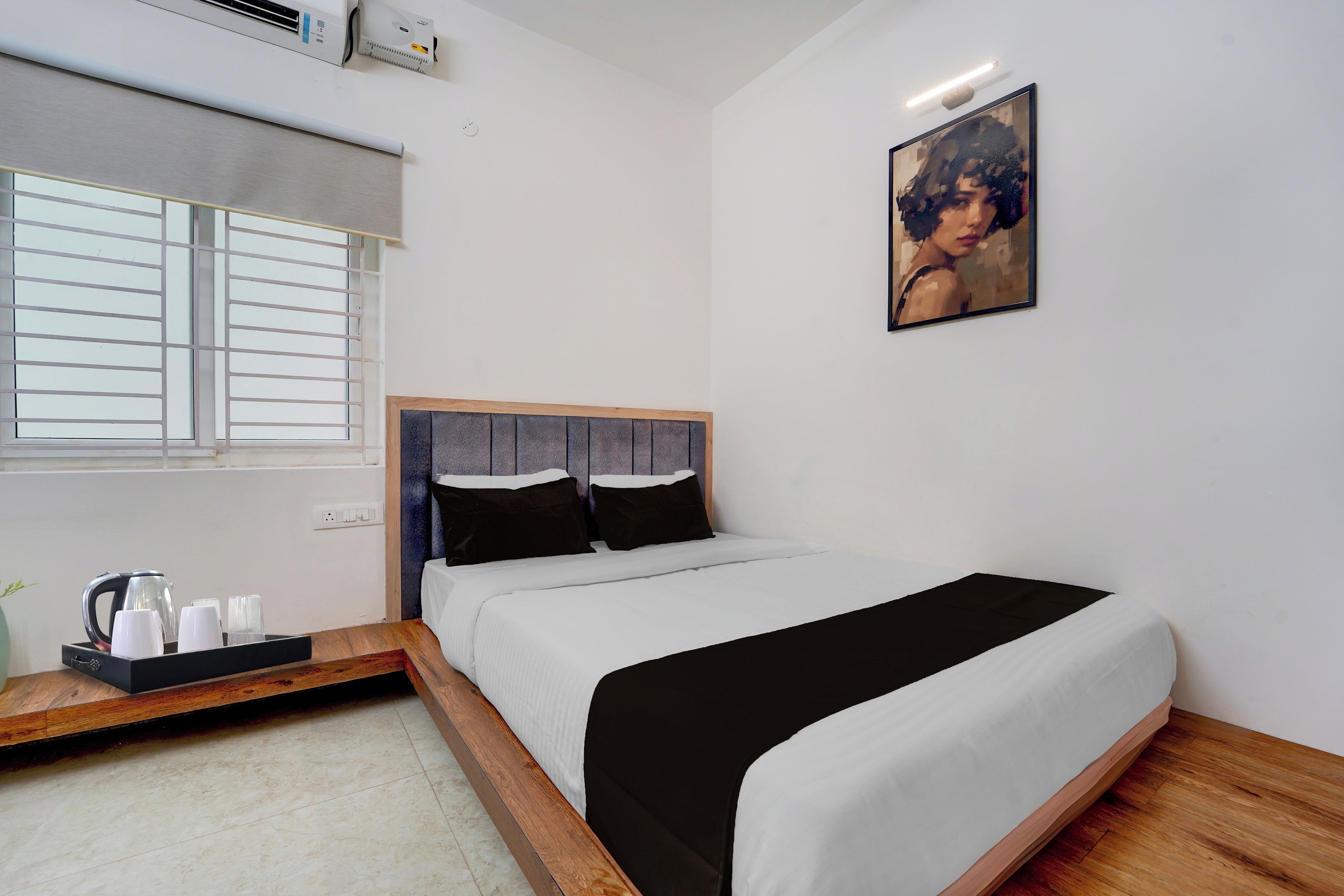 Un dormitorio con cama, ventana con persianas, un cuadro en la pared y una mesita de noche con tetera y tazas.
