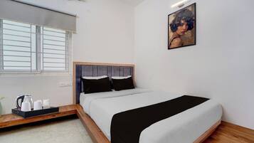 Un dormitorio con cama, ventana con persianas, un cuadro en la pared y una mesita de noche con tetera y tazas.