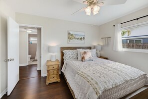 Un dormitorio con cama, mesitas de noche, ventilador de techo, ventana con vistas y puerta que da a un baño.