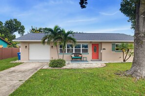 A single-story house with a garage, a red door, and a small porch with a bench. - Pet Friendly 2BR Fenced-In near St. Pete Beach (St. Petersburg)