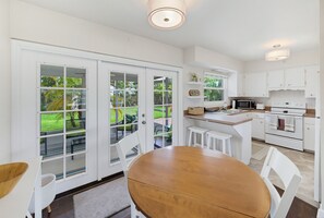 Una cocina moderna con muebles blancos, un comedor con mesa y sillas de madera, y vista a un jardín verde a través de amplios ventanales.