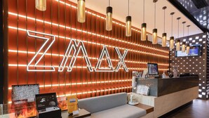 A reception area with a modern design, featuring a large illuminated 'ZMAX' sign, a reception desk, seating area, and a digital screen displaying information. - Zmax Hotels (Shenzhen)