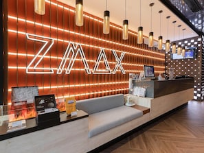 A reception area with a modern design, featuring a large illuminated 'ZMAX' sign, a reception desk, seating area, and a digital screen displaying information. - Zmax Hotels (Shenzhen)