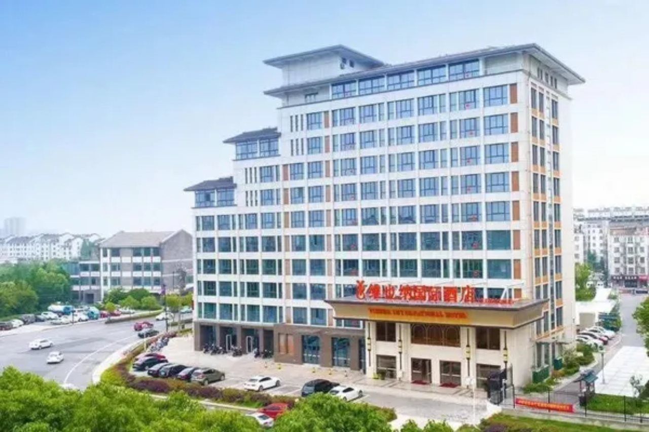 A multi-story hotel with a modern facade, a parking lot, and a clear sky.