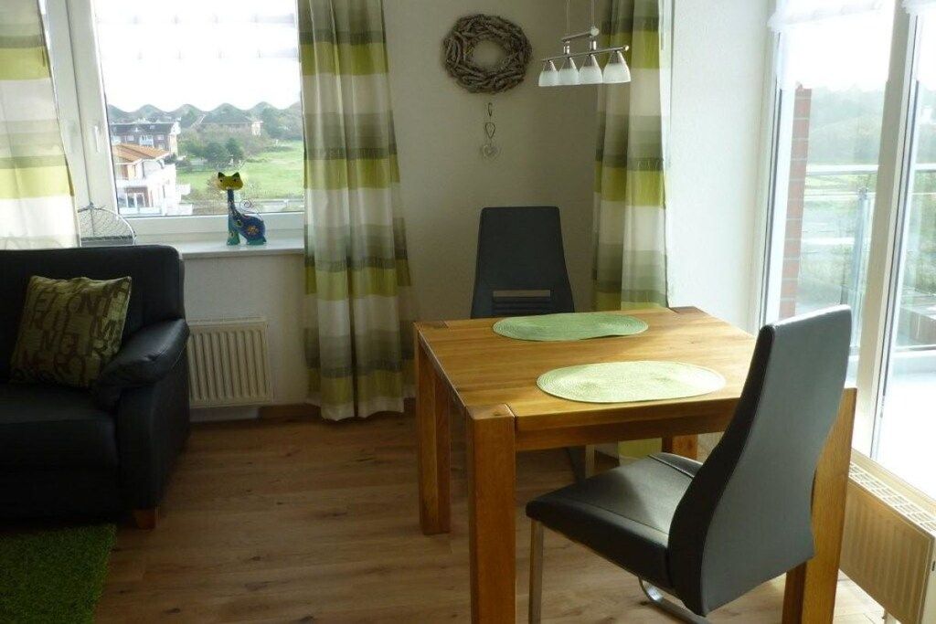 A room with a wooden dining table, a black chair, a black sofa with a cushion, a window with curtains, and a view of houses outside.