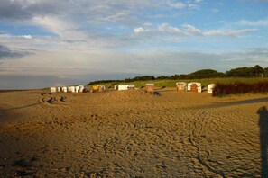 A beach with several beach huts aligned in a row, a sandy path leading to them, and a grassy area with trees in the background. - Holiday apartment with 1 bedroom (Cuxhaven-Sahlenburg)