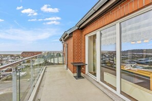 Een balkon met glazen hekwerk met uitzicht op een strand en een parkeerplaats.