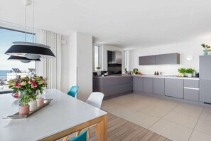 A modern kitchen with grey cabinets, a dining area with a white table, and a large window overlooking the sea. - Penthouse on the Baltic Sea (Kappeln)