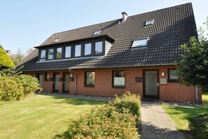 A two-story brick house with a sloped roof, a front door, and a small front yard with a lawn and shrubs. - Holiday apartment with 1 bedroom (St. Peter-Ording)