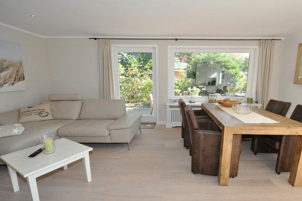 Una sala con un sofá, una mesa de centro y una mesa de comedor con sillas. Hay una ventana con cortinas y vistas a zonas verdes.