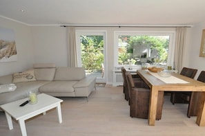 A living room with a sofa, a coffee table, and a dining table with chairs. There is a window with curtains, and a view of greenery outside. - Holiday apartment with 1 bedroom (St. Peter-Ording)