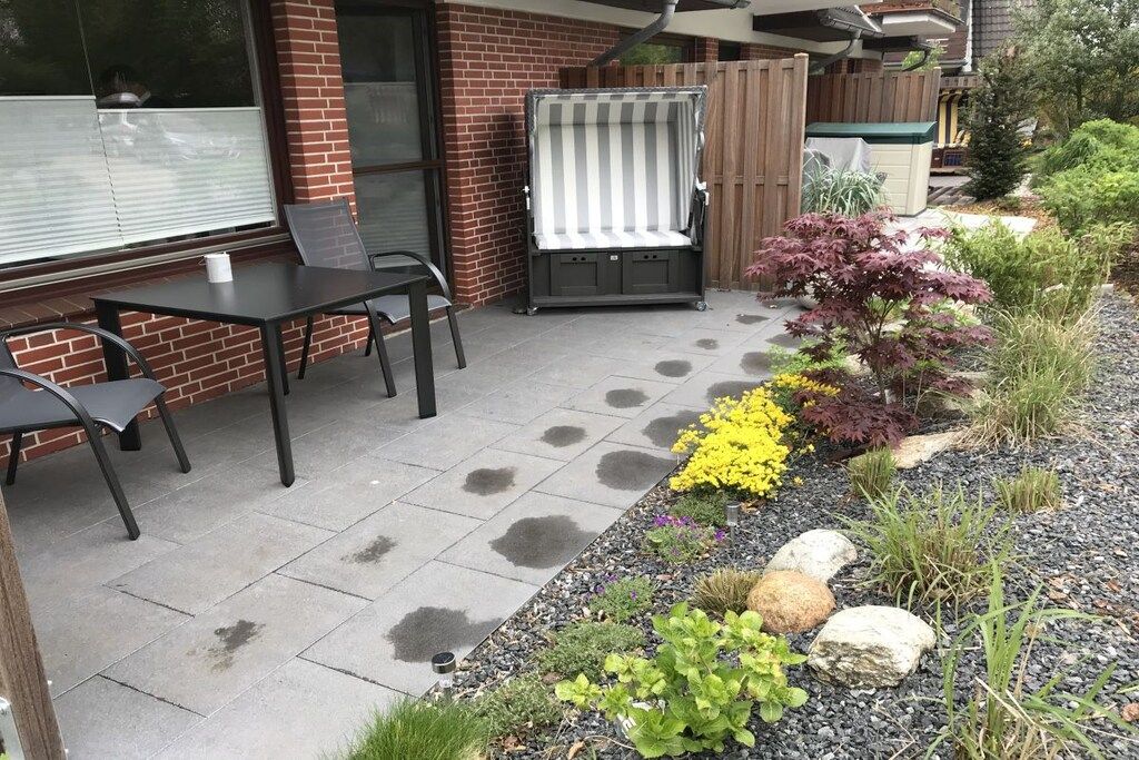 Een terras met een tafel en stoelen, een overdekte zithoek, en een tuin met verschillende planten en stenen.
