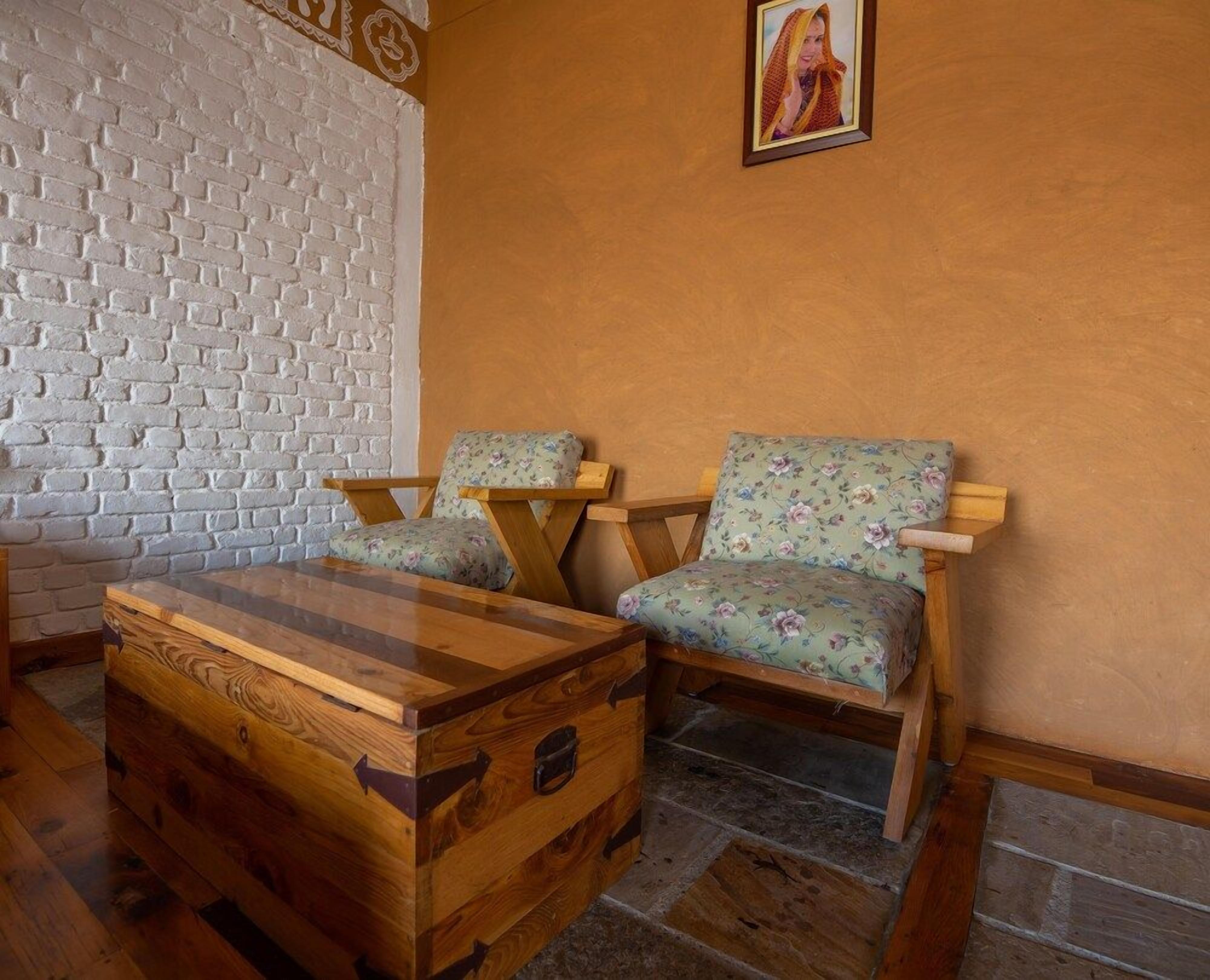 A room with a white brick wall, a wooden chest, two floral-patterned chairs, and a framed picture on the wall.