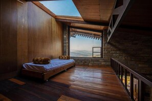 A room with a sloped ceiling, a bed, a large window, and a stone wall. - Blusalzz Homes - The Himalayan Bungalow, Almora - Uttarakhand by Zuzu (Almora)