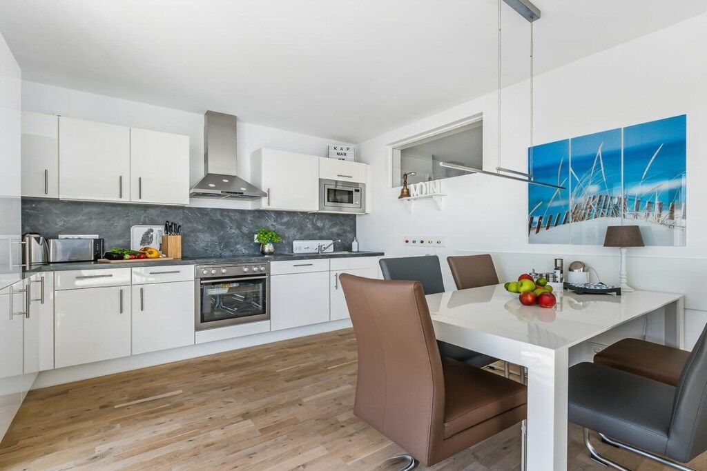 A modern kitchen with white cabinets, a marble countertop, and a dining area with a white table and brown chairs.