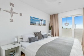 Ein Schlafzimmer mit einem Bett, einem Nachttisch, einer Lampe, einem Wandkompass, einem gerahmten Bild und einem Fenster mit Vorhängen.