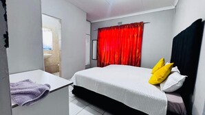A bedroom with a bed, a window with red curtains, a white wall, and a door leading to a bathroom. - Charming 2-bedroom estate with fitness room in Woodland Hills Estate (Woodland Hills Estate, Bloemfontein)