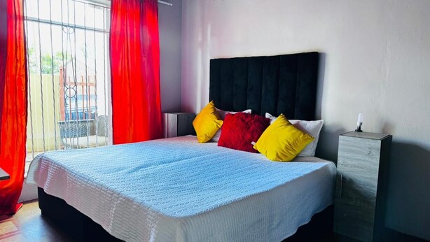 A bed with a white quilt, a black headboard, and multiple pillows in red and yellow. - Charming 2-bedroom estate with fitness room in Woodland Hills Estate (Woodland Hills Estate, Bloemfontein)