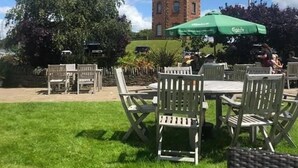 An outdoor seating area with tables and chairs, a green umbrella, and a brick tower in the background. - Towers Hotel & Spa (Neath)