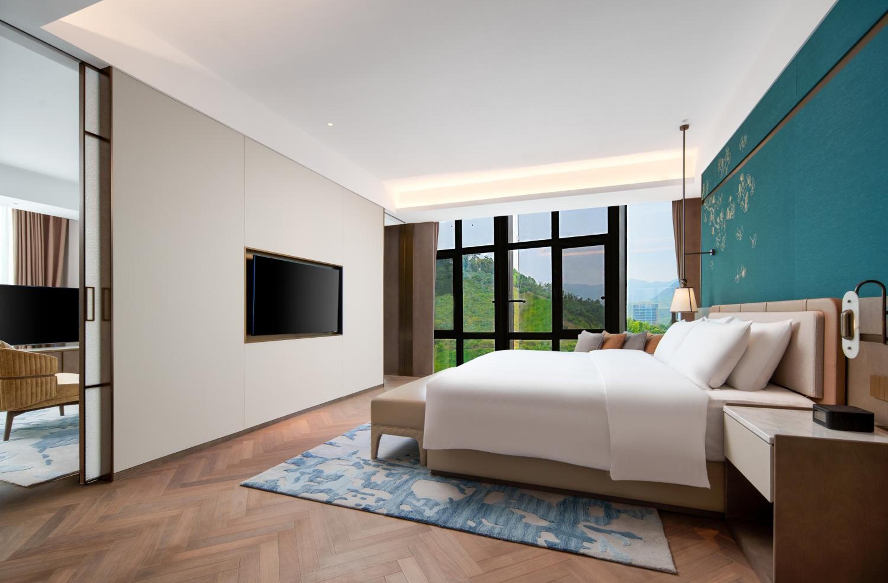 A modern hotel room with a large bed, a flat-screen TV, a desk, and a view of the outdoors.