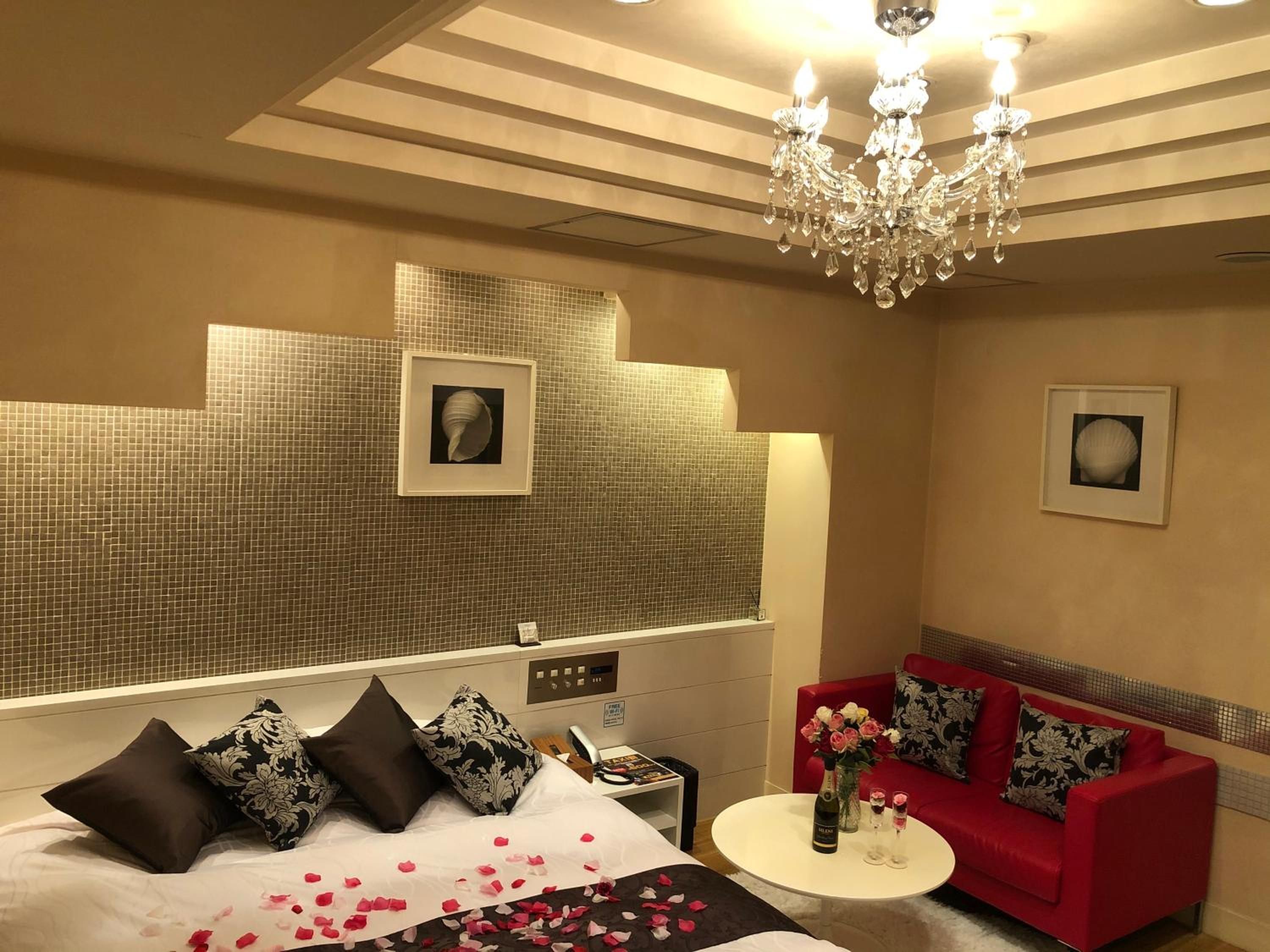 Quarto de hotel com cama, sofá vermelho, pequena mesa com garrafa e copos, e um lustre.