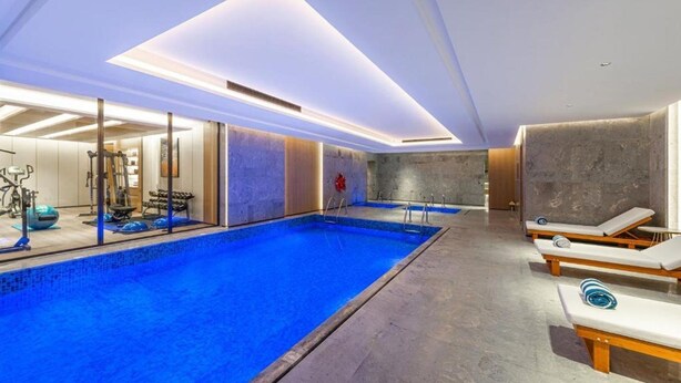 Una piscina interior moderna con superficie de agua cristalina, rodeada de sillones de descanso y equipo de ejercicio.