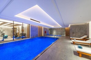 Una piscina interior moderna con superficie de agua cristalina, rodeada de sillones de descanso y equipo de ejercicio.