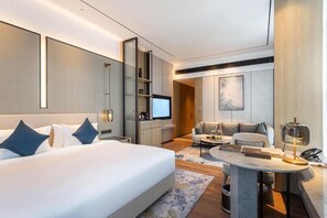 A modern hotel room with a large bed, a desk with a lamp, a television, and a sofa.