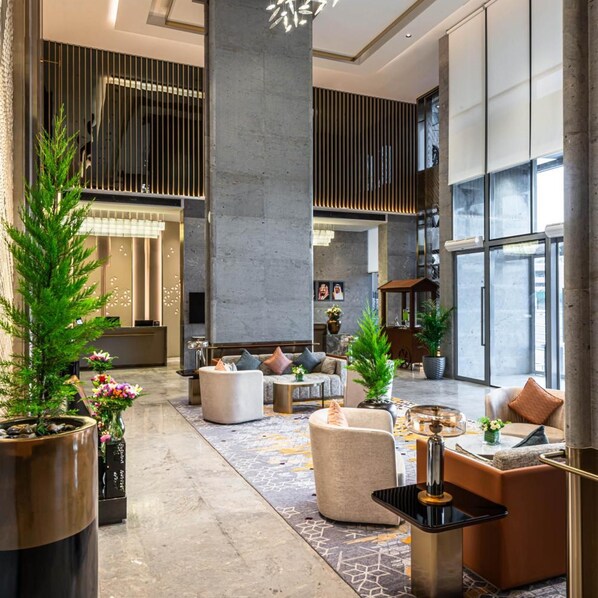 A spacious lobby with a modern design, featuring a central seating area with sofas and armchairs, a large pillar, and a variety of potted plants.