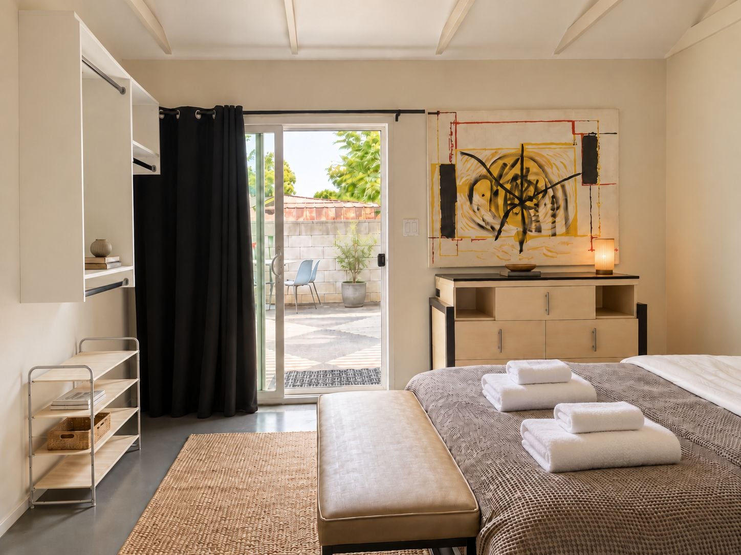 A hotel room with a bed, a desk, a chair, a bench, and a large window with a view of the outside.