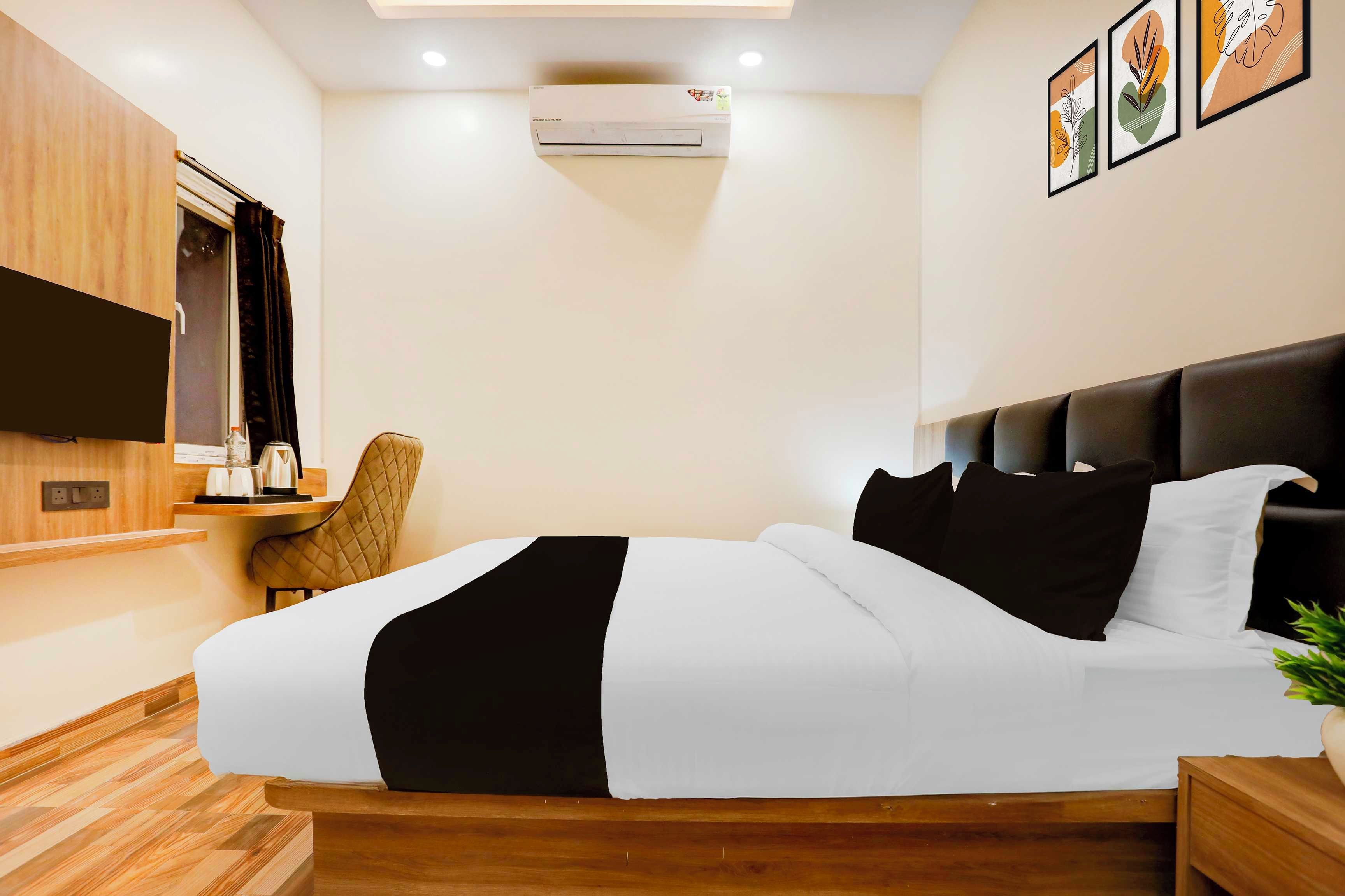 Habitación de hotel con cama, televisor de pantalla plana en la pared, mesita de noche con lámpara, una silla y planta en maceta.
