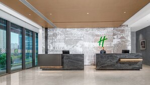 A modern reception area with a large digital screen displaying a green character, two reception desks, and a view of a cityscape through large windows. - Holiday Inn Kunshan East (Suzhou)
