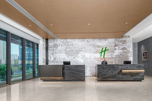 A modern reception area with a large digital screen displaying a green character, two reception desks, and a view of a cityscape through large windows. - Holiday Inn Kunshan East (Suzhou)