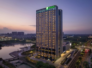 A tall, modern hotel with a grid of windows, a Holiday Inn sign, and a view of a cityscape and river at dusk. - Holiday Inn Kunshan East (Suzhou)