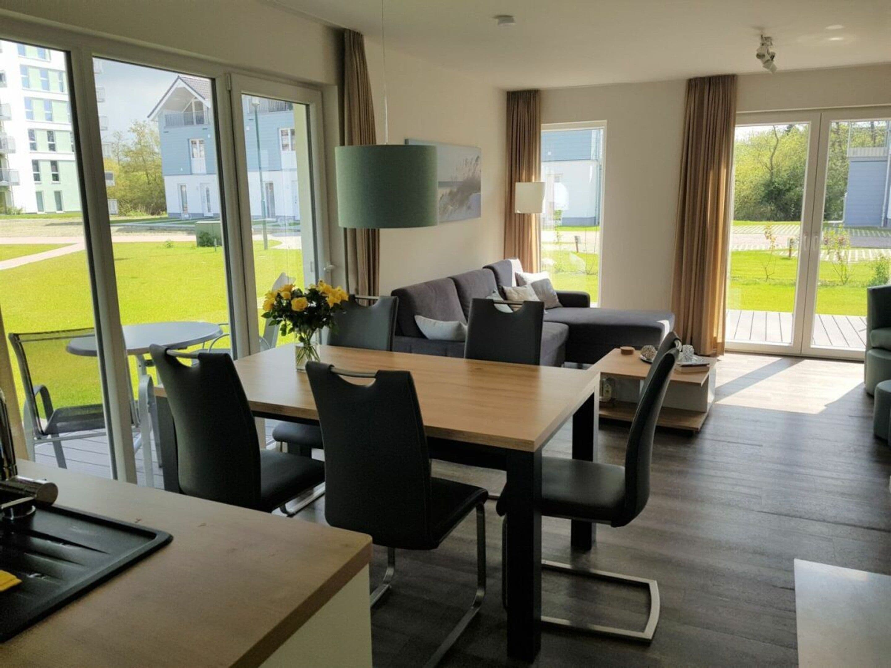 Een moderne woonkamer met een eethoek, grote ramen en uitzicht op een gebouw en groen buiten.