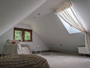 A room with a sloped ceiling, a window with a view of greenery, a white chair, a small table, and a woven ottoman.