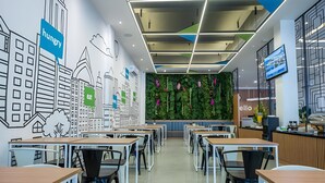 Un comedor moderno con mesas y sillas, una pared verde con flores y un mural de un paisaje urbano.