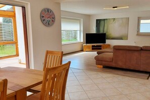 A living room with a wooden dining table, a brown sofa, a television on a wooden stand, and a painting on the wall. - Holiday apartment with 4 bedrooms (Cuxhaven)