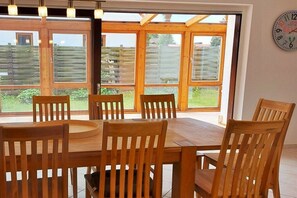 A dining area with a wooden table and chairs, large windows with a view of a garden, and a wall clock. - Holiday apartment with 4 bedrooms (Cuxhaven)