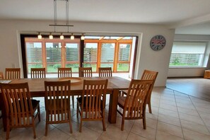 A dining area with a large wooden table and chairs, large windows with a view of a garden, and a wall clock. - Holiday apartment with 4 bedrooms (Cuxhaven)