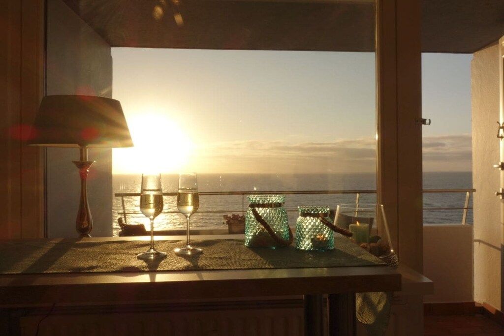 A table with a glass of wine, two glass jars, and a candle, overlooking a sunset over the sea.