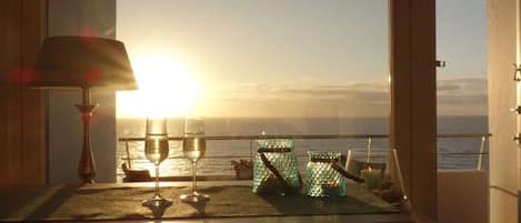 A table with a glass of wine, two glass jars, and a candle, overlooking a sunset over the sea.
