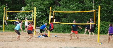 Eine Gruppe von Menschen spielt Beachvolleyball auf einem Sandplatz, im Hintergrund sind Bäume zu sehen.