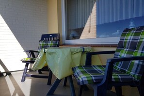 Eine Terrasse mit Tisch und zwei Stühlen, ein Fenster mit Jalousien und eine Backsteinmauer.