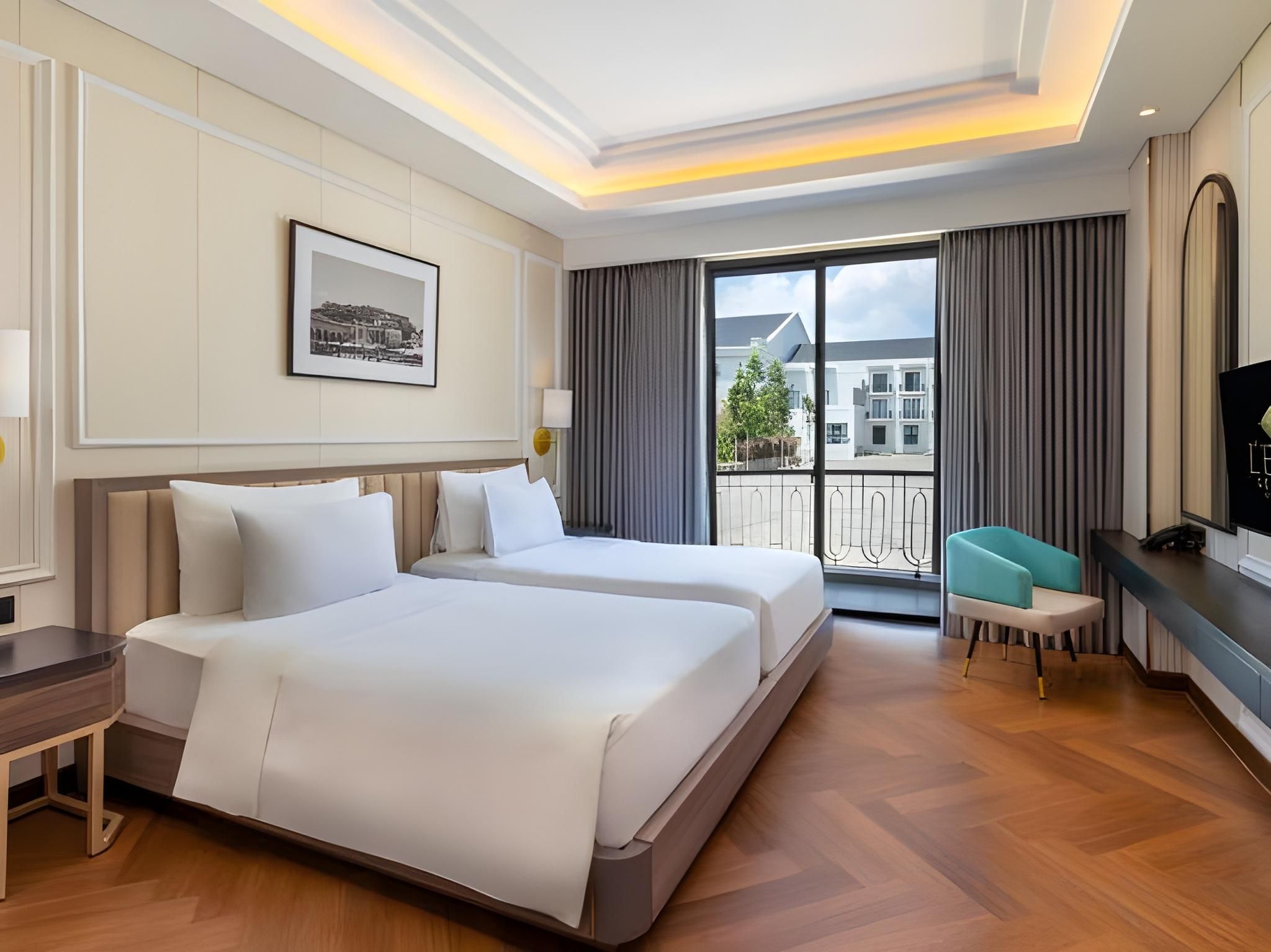 Habitación de hotel con dos camas, un escritorio, una silla, un televisor y un ventanal con vistas a edificios.