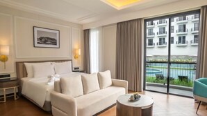 Een hotelkamer met een bed, een bank, een ronde salontafel en uitzicht op een gebouw en zwembad.